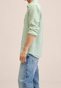 Chemise vert clair à manches longues en tissu texturé, portée avec un jean bleu. La chemise est légèrement froissée avec un détail de poche.
