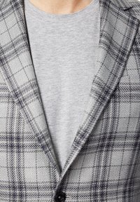 Szary marynarka w kratę z teksturowanej wełny, z klapami oraz zapięciem na jeden guzik, noszona na jasnoszarej koszuli.