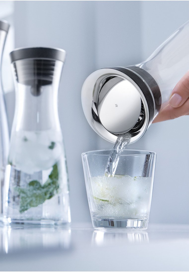 Carafe en verre versant de l'eau dans un verre transparent avec des glaçons, des feuilles de menthe visibles. La carafe a un couvercle en acier inoxydable élégant et un design courbé.