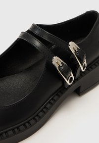 Black leather shoes with two straps, featuring silver buckles. The sole has a textured, rugged design for added grip.