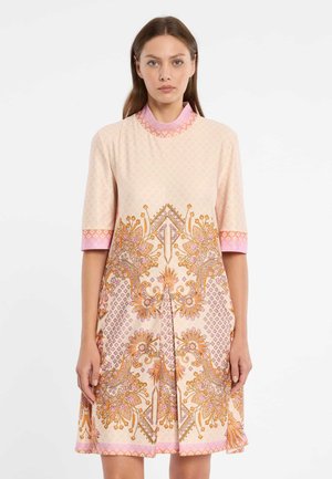 Femme portant une robe crème jusqu'aux genoux avec des motifs floraux et géométriques rose et orange, debout devant un fond blanc.