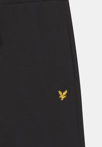 Svart tyg med en liten gul broderad fågel-logotyp nära sömmen.