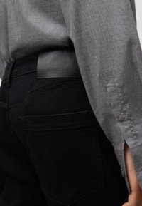 Camicia button-up grigia, jeans di denim neri con un'etichetta in pelle strutturata. Inquadratura ravvicinata del polsino della camicia e dei dettagli della vita.