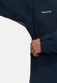 Veste imperméable navy avec une poche zippée, des poignets ajustables et une texture lisse ; présente le nom de la marque "Mammut" sur la poitrine.