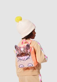 Kind met krullend haar die een lichtroze muts met een gele pompon draagt, terwijl hij een tas met kattenmotief en kleurrijke accenten draagt.