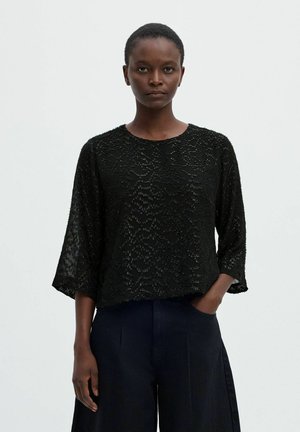 Le modèle se tient devant un fond uni, vêtu d’un chemisier noir à texture marquée et à manches trois-quarts, ainsi que d’un pantalon foncé à jambes larges.
