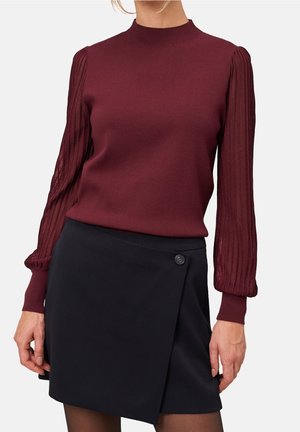 Woman wearing a burgundy long-sleeve top with pleated sleeves and a black wrap skirt with a single button closure.