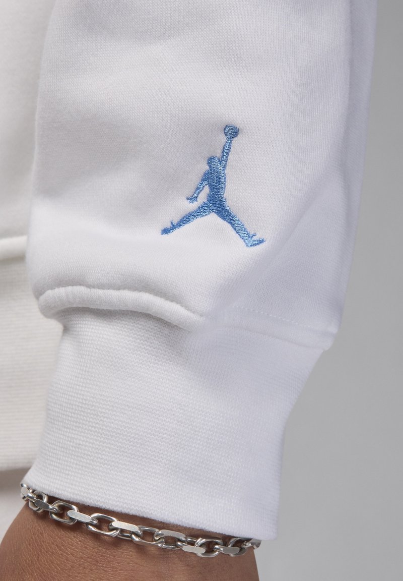 Sweatshirt blanc avec un logo Jumpman brodé bleu sur la manche. Il présente des poignets côtelés et un bracelet en chaîne argenté au poignet.