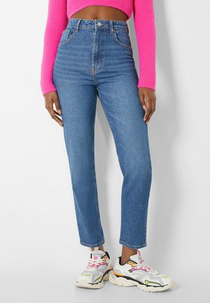 Personne portant un jean taille haute bleu, des baskets blanches avec des accents colorés et un pull court rose vif, debout devant un fond blanc.