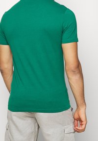 Man med grön kortärmad t-shirt och ljusbeige cargoshorts, stående mot en enkel vit bakgrund, sedd bakifrån.