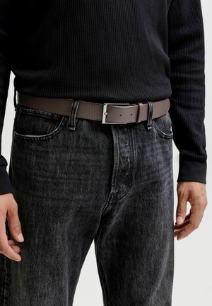 Mees kannab musta soonikutega kampsunit, musta pestud teksapükse ja pruuni nahast vööd ristküliku kujulise hõbedase pandlaga, käed lõdvestunult külgedel.