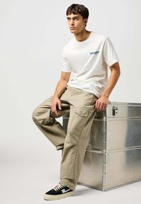 Biała koszulka z krótkim rękawem z niebieskim logo, beżowe spodnie cargo z kieszeniami i czarne sneakersy niskie z kremowymi akcentami, siedzący na metalowej skrzynce.