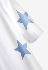 T-shirt bianca in cotone con stampe di stelle blu sulle maniche. Materiale morbido, maniche corte, scollo rotondo, con una finitura leggermente testurizzata.