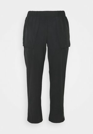 Persona que lleva pantalones negros recortados con cintura elástica y grandes bolsillos laterales, combinados con zapatillas blancas y una camiseta blanca.
