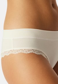 Culotte en coton beige avec une taille douce et extensible et une délicate bordure en dentelle le long des ouvertures de jambe, présentant une texture lisse.