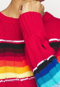 Pull en tricot présentant de larges rayures horizontales rouges, orange, jaunes, marron, noires et bleues, avec un petit logo sur la manche.