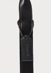 Brățară din piele neagră cu o textură netedă, având un tag rectangular atașat printr-un lanț, embosat cu textul "COACH".