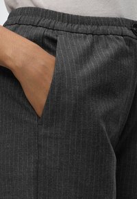Main placée dans la poche avant d’un pantalon gris foncé à fines rayures, avec une ceinture élastique et une fermeture à bouton.