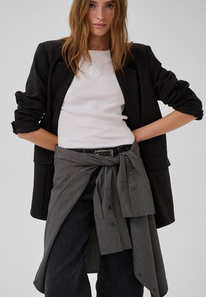 Donna che indossa una maglietta bianca, blazer nero con maniche arrotolate, jeans neri e una camicia a righe legata in vita, in piedi con le mani sui fianchi.