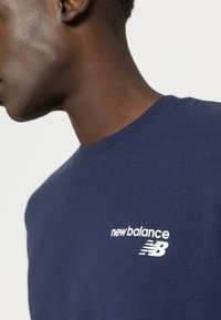 Námořnické modré bavlněné tričko s bílým logem a grafikou "New Balance" na hrudi, klasický kulatý výstřih a krátké rukávy.