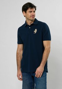 Tamno plava polo majica od pamuka, s kragnom i kratkim rukavima. Uključuje svijetlu izvezenu dekoraciju na lijevoj strani grudi.