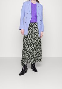 Femme portant un blazer double boutonnage lavande, un haut violet, une longue jupe fleurie et des bottines à talons noires sur fond blanc.