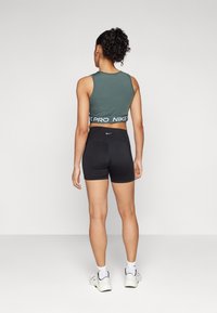 Zelený sportovní crop top s bílým logem Nike na pásku, kombinovaný s černými přiléhavými šortkami. Modelka je otočená zády, zobrazující zadní design.