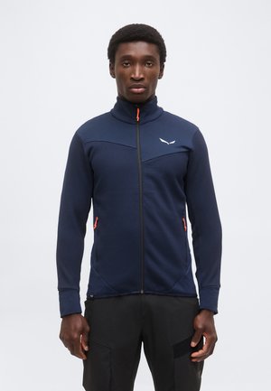 Mężczyzna w granatowej teksturowanej kurtce na zamek błyskawiczny z jaskrawo-pomarańczowymi suwakami i czarnych spodniach, stojący na jednolitym jasnym tle.