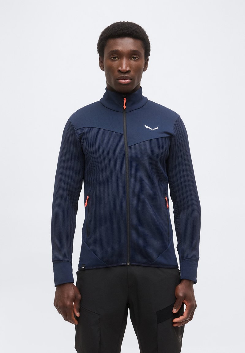 Άνδρας που φοράει σκούρο μπλε υφασμάτινο μπουφάν με φερμουάρ και λαμπερά πορτοκαλί τσεπετζήδες φερμουάρ και μαύρο παντελόνι, που στέκεται μπροστά σε ένα απλό φωτεινό φόντο.