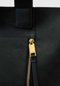Sac fourre-tout en cuir noir à texture lisse, équipé d'une fermeture éclair dorée et d'une languette en cuir. Comprend une pièce cousue sur la poignée.