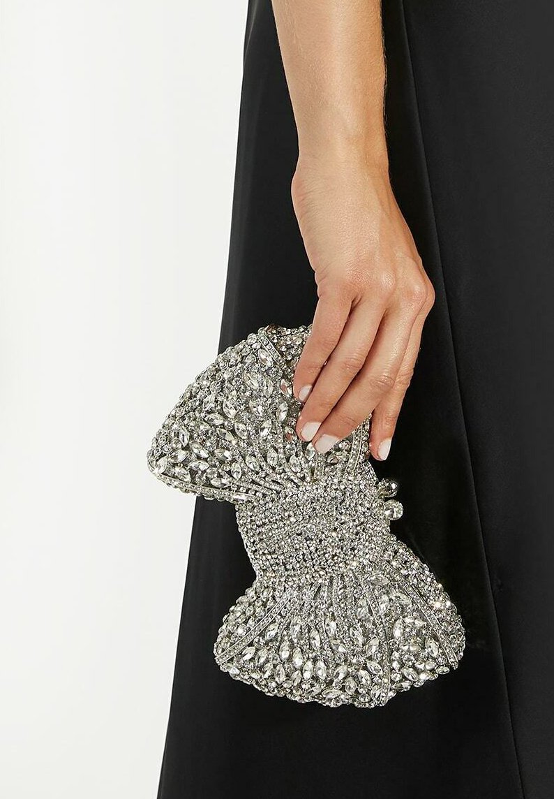 Hand holding a silver, gem-encrusted, bow-shaped clutch bag against a black dress background.