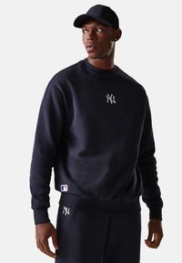 Hombre con sudadera negra de los New York Yankees, pantalones a juego y gorra negra, mirando hacia un lado sobre un fondo claro y liso.