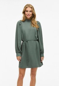 Groene jurk met riem, lange mouwen en geplooide schouderdetails. Gemaakt van getextureerde stof met een hoge hals en een ontspannen silhouet.