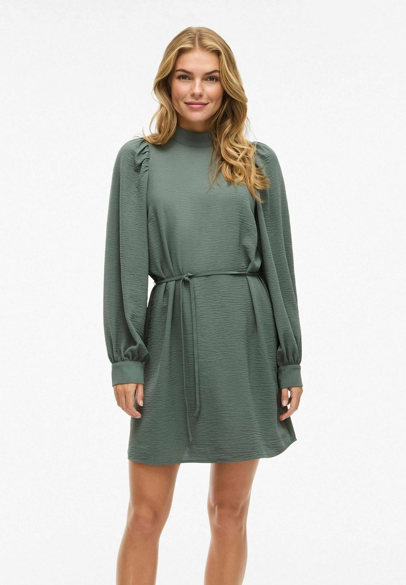 Groene jurk met riem, lange mouwen en geplooide schouderdetails. Gemaakt van getextureerde stof met een hoge hals en een ontspannen silhouet.
