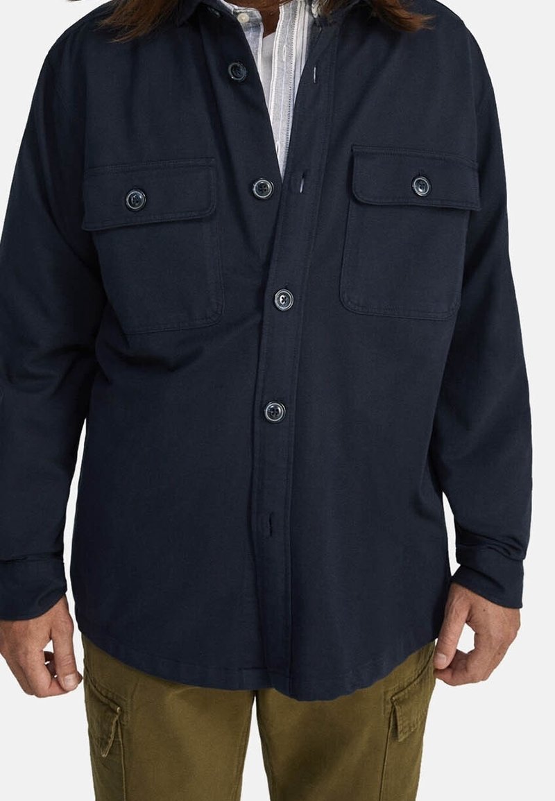 En man som bär en mörk marinblå knappad skjorta med två bröstfickor över en delvis synlig randig skjorta, parat med olivgröna cargo-byxor.