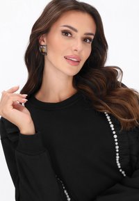 Fekete pulóver fodros ujjakkal és gyöngy díszítéssel az oldalán. A modell vastag, négyzet alakú fülbevalót visel, és hosszú, hullámos hajat mutat.