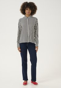 Camicia a righe bianca e nera, abbinata a jeans blu e ballerine rosse. Caratteristiche notevoli includono un colletto a punta e maniche lunghe.
