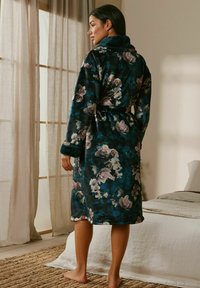 Woman standing barefoot in a floral dark blue robe with a belt, facing a window with sheer curtains in a softly lit bedroom.