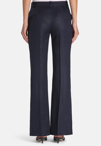 Femme portant un pantalon large bleu marine foncé avec passants pour ceinture, poches arrière, et sandales à talons noires ouvertes à l'avant, debout devant un fond blanc.