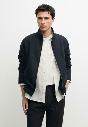 Giovane uomo con giacca nera con zip sopra una camicia bianca con bottoni e pantaloni scuri, in piedi davanti a uno sfondo bianco semplice.