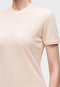 Chemise rose clair texturée à manches courtes avec bords festonnés, petit logo brodé sur la poitrine gauche, portée par une personne visible du cou à la taille.