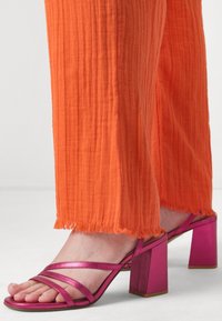 Sandales à talons hauts roses à lanières avec un talon carré, associées à un pantalon orange texturé à bords effilochés. Gros plan sur les chaussures et le détail de l'ourlet.