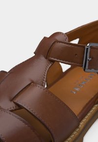 Καφέ δερμάτινο σανδάλι με πολλαπλούς ιμάντες και ασημί αγκράφα, που δείχνει λεπτομερή ραφή και επενδεδυμένο με μαξιλαρωτό πάτο.