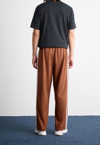 Braune Jogginghosen mit weißen Seitestreifen, kombiniert mit einem schwarzen T-Shirt mit kurzen Ärmeln. Der Stoff wirkt weich; lockere Passform mit elastischem Bund.