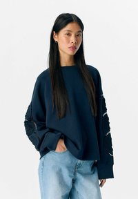 Marineblauwe sweatshirt met lange mouwen, versierd met bloemenkantaccenten, ontspannen pasvorm en een ronde hals. Gecombineerd met lichtblauwe denim jeans.