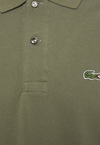 Olive zelené polo tričko vyrobené z textilného materiálu, s klasickým golierom, dvoma gombíkmi a vyšitým logom krokodíla na hrudi.