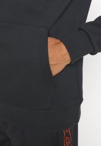 Zwarte sweatshirt met een kangoeroezak, ribgebreide manchetten en zoom. Heeft een oranje accent op de bijbehorende broek. Soepele textuur.