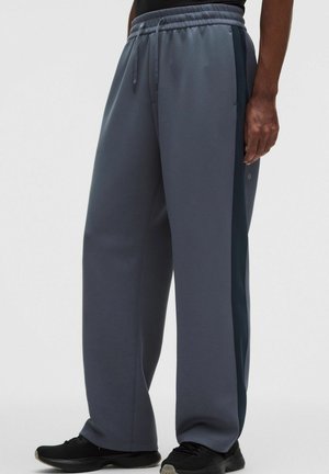 Pantalones deportivos grises de corte holgado con cintura elástica y cordón, usados con zapatillas deportivas negras, mostrados desde la mitad del torso hasta los pies.
