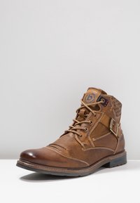 Stivaletto in pelle marrone con punta rotonda, superficie testurizzata, cuciture a contrasto, design con lacci e un dettaglio decorativo a fibbia laterale.