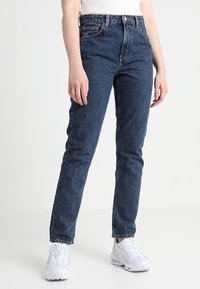 Jeans de mezclilla azul de talle alto con piernas rectas, que presentan un diseño clásico de cinco bolsillos y detalles de costura sutiles, combinados con zapatillas blancas.